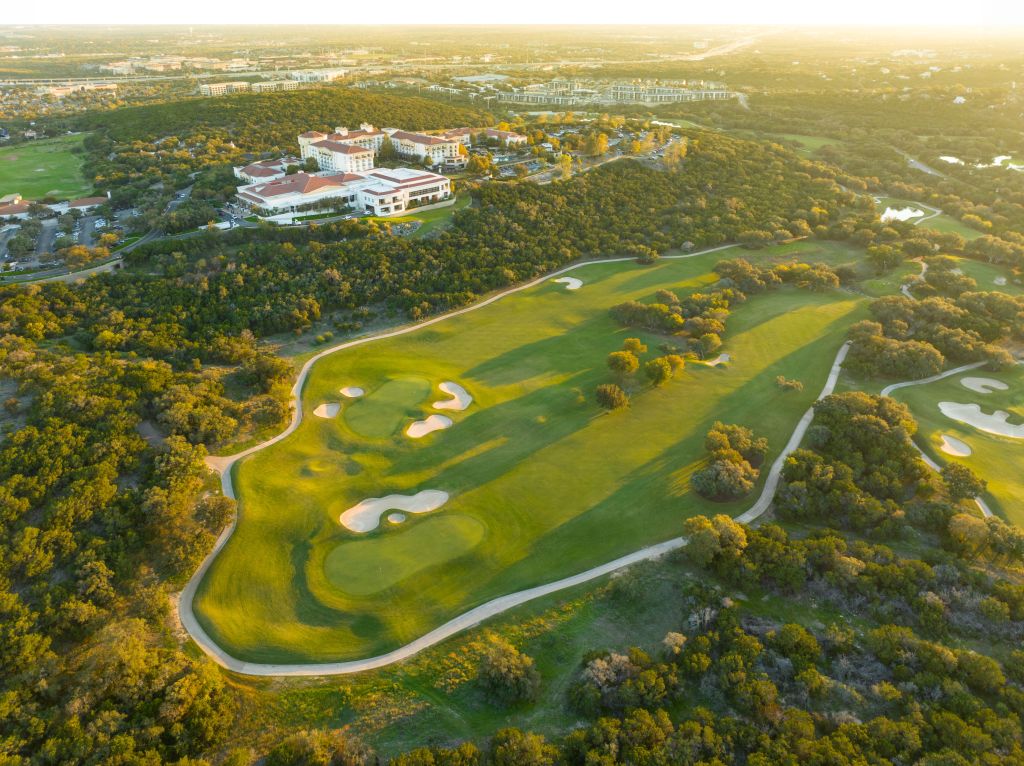 A birds eye view of the golf course with the clubhouse near the top left. Surrounded by trees on a sunny day.
